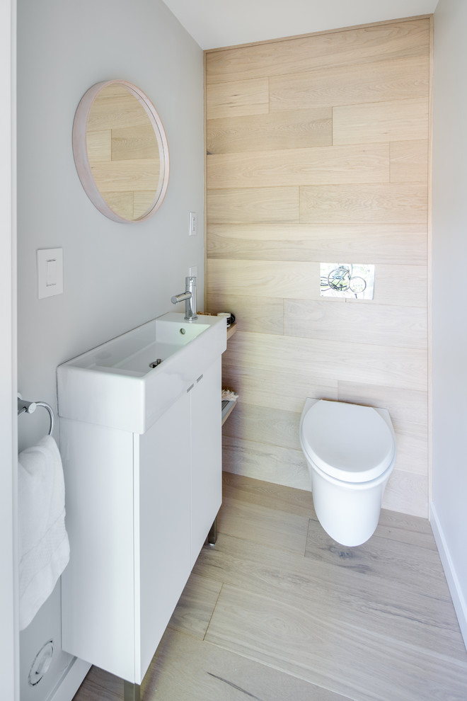 Inredning av ett modernt litet toalett, med släta luckor, vita skåp, en vägghängd toalettstol, vita väggar, ljust trägolv, ett konsol handfat och vitt golv