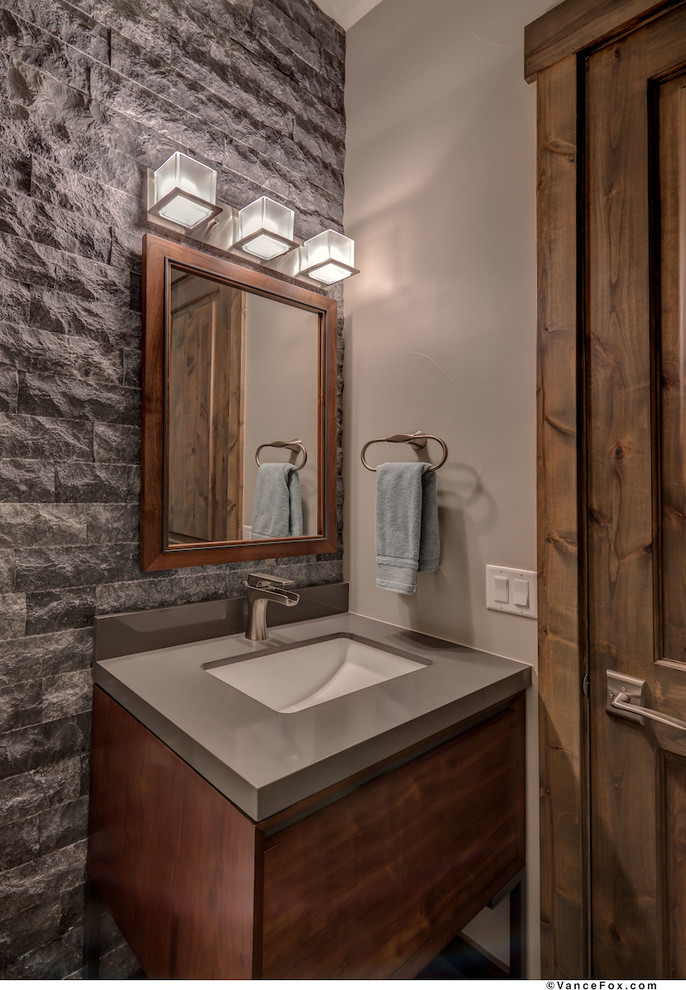 Inredning av ett klassiskt litet grå grått toalett, med möbel-liknande, bruna skåp, en toalettstol med separat cisternkåpa, grå kakel, stenhäll, grå väggar, ett undermonterad handfat och bänkskiva i akrylsten