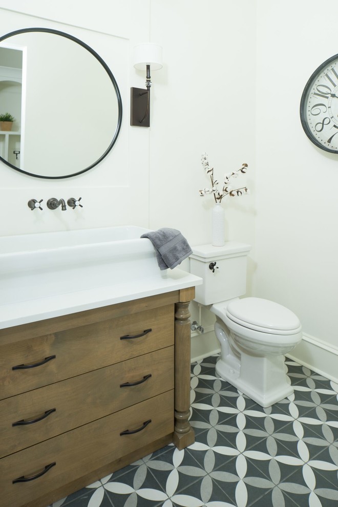 Example of a mid-sized farmhouse ceramic tile and multicolored floor powder room design in Boise with furniture-like cabinets, medium tone wood cabinets, a two-piece toilet, white walls, a trough sink and white countertops