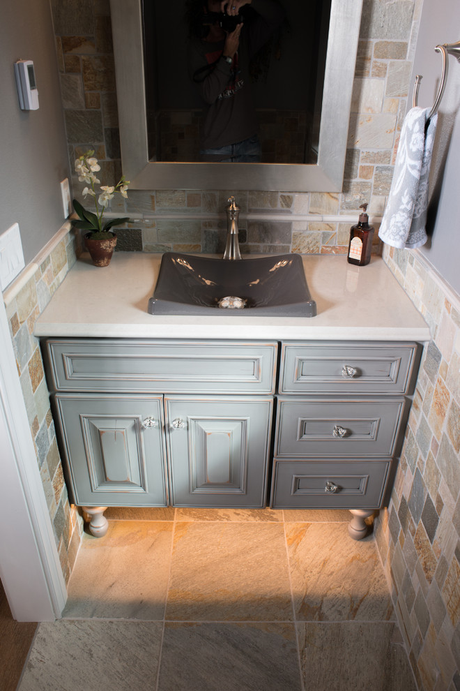 Example of a mid-sized mountain style multicolored tile and stone tile slate floor powder room design in Other with raised-panel cabinets, a two-piece toilet, gray walls, a vessel sink, granite countertops and distressed cabinets
