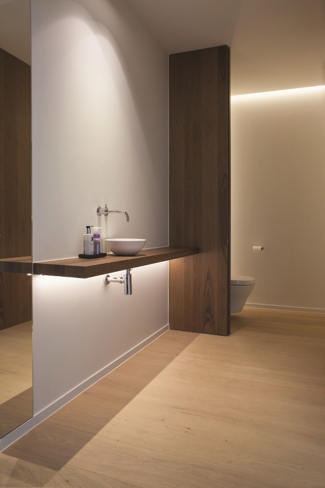 Mid-sized minimalist light wood floor powder room photo in Dallas with a vessel sink, wood countertops, a wall-mount toilet, white walls and brown countertops