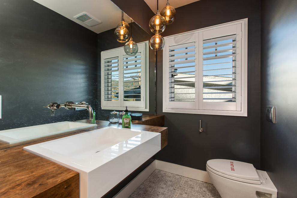 Small transitional cement tile floor and gray floor powder room photo in Los Angeles with open cabinets, a wall-mount toilet, black walls, a drop-in sink, wood countertops and brown countertops