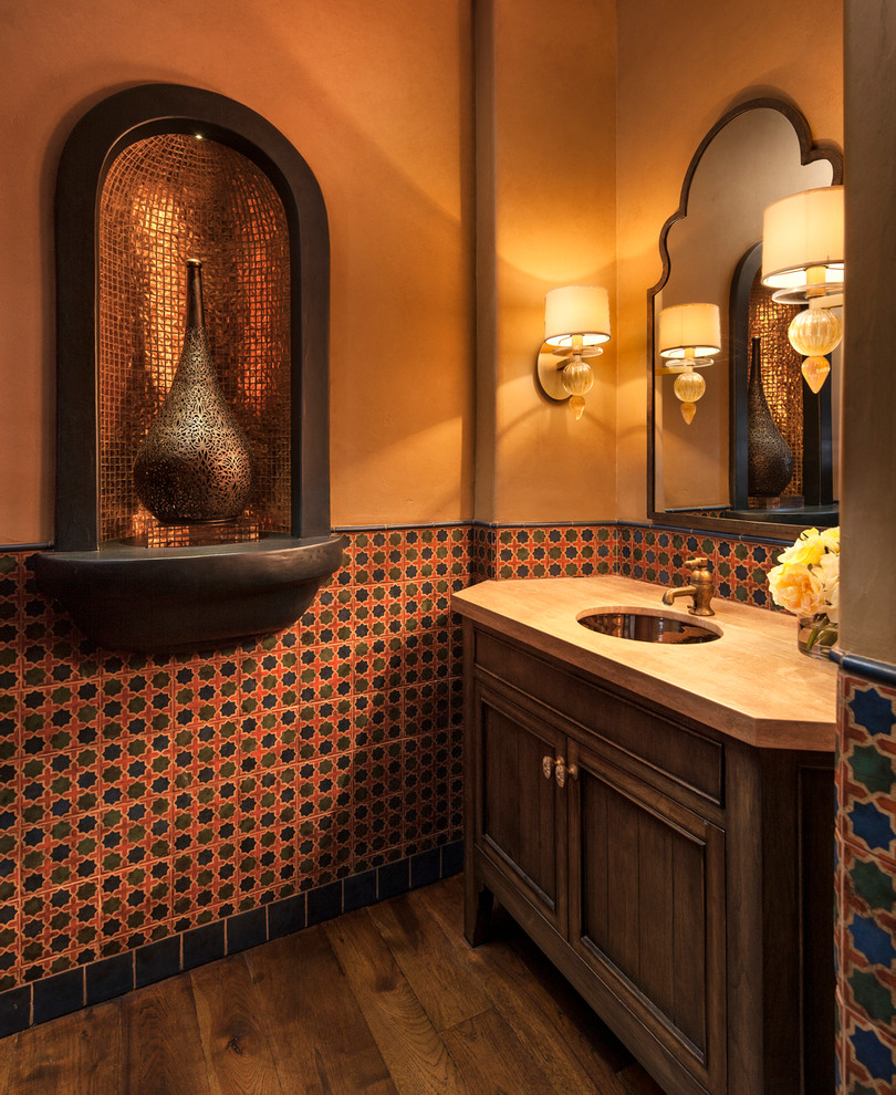 サンタバーバラにある地中海スタイルのおしゃれなトイレ・洗面所 (アンダーカウンター洗面器、家具調キャビネット、濃色木目調キャビネット、マルチカラーのタイル、オレンジの壁、濃色無垢フローリング、ベージュのカウンター) の写真