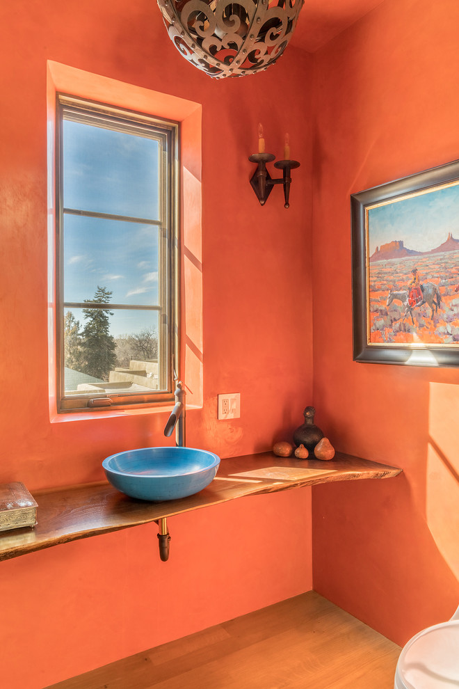 アルバカーキにあるサンタフェスタイルのおしゃれなトイレ・洗面所 (オレンジの壁、無垢フローリング、ベッセル式洗面器、木製洗面台、ベージュの床、ブラウンの洗面カウンター) の写真