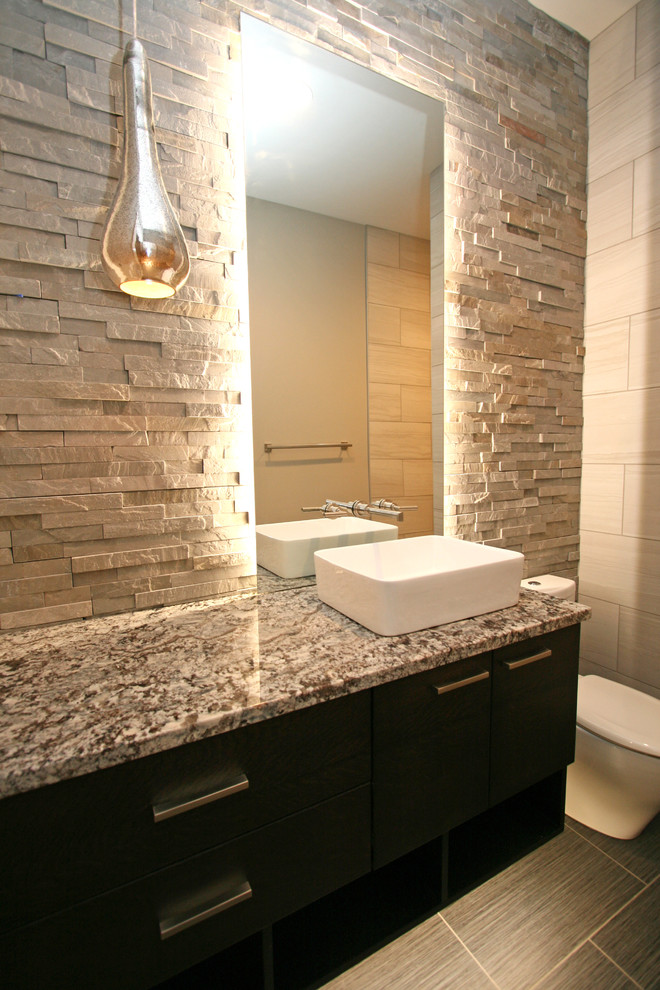 Bild på ett mellanstort funkis toalett, med ett fristående handfat, släta luckor, svarta skåp, granitbänkskiva, en toalettstol med hel cisternkåpa, grå kakel, stenkakel, grå väggar och klinkergolv i porslin