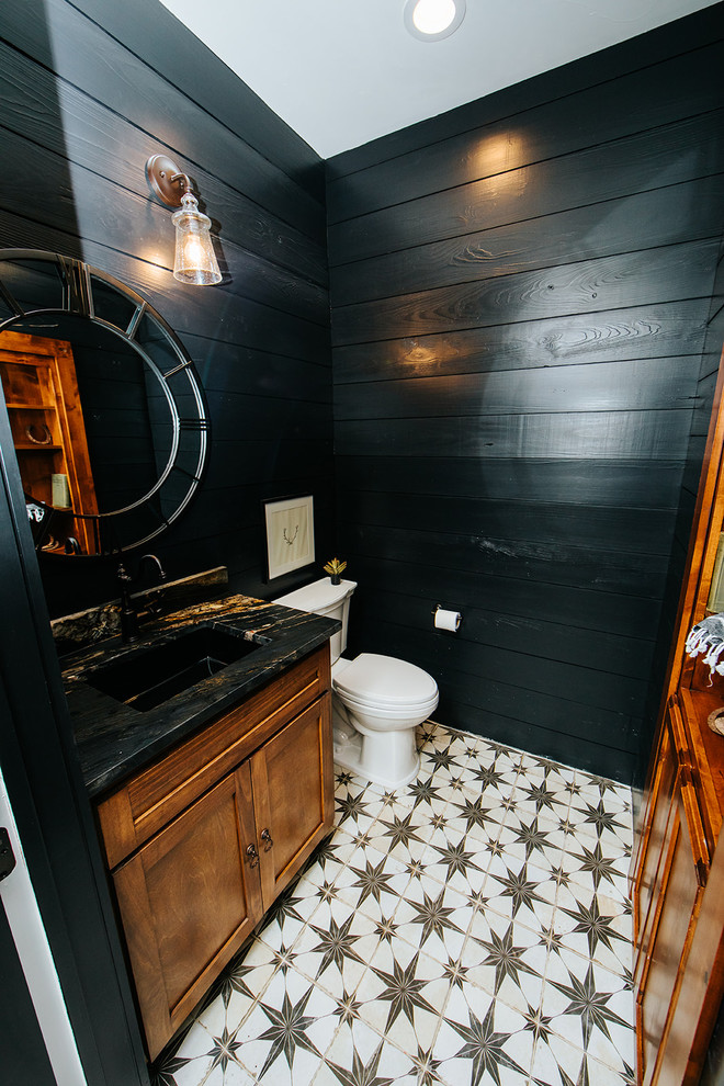 Bild på ett litet lantligt svart svart toalett, med skåp i shakerstil, bruna skåp, en toalettstol med hel cisternkåpa, svarta väggar, cementgolv, ett nedsänkt handfat och granitbänkskiva
