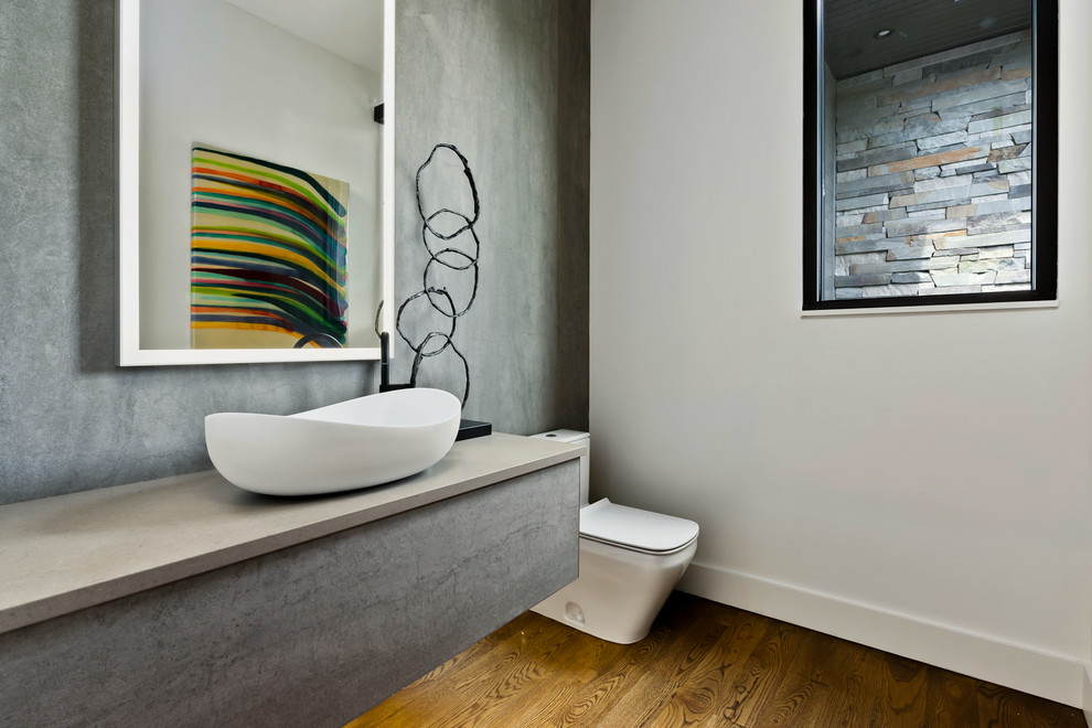 Modelo de aseo actual con sanitario de una pieza, paredes grises, suelo de madera en tonos medios, lavabo sobreencimera y encimeras grises