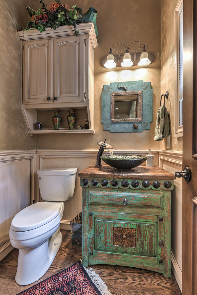 Photo of a medium sized cloakroom in Oklahoma City with recessed-panel cabinets, distressed cabinets, a two-piece toilet, beige walls, dark hardwood flooring, a vessel sink, wooden worktops and brown floors.