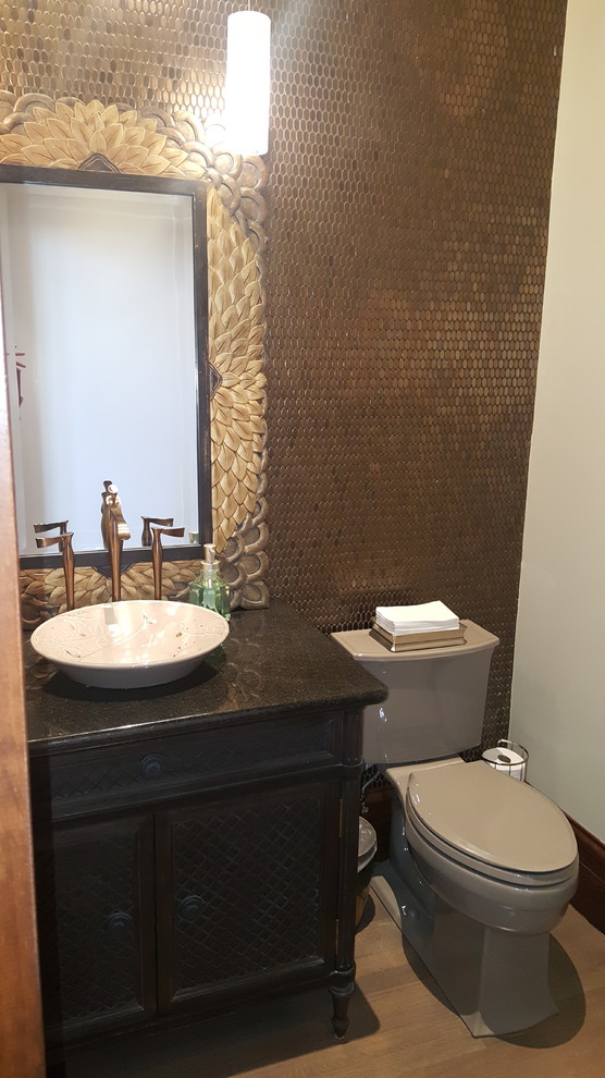 Klassisk inredning av ett stort toalett, med möbel-liknande, svarta skåp, en toalettstol med separat cisternkåpa, kakel i metall, bruna väggar, ljust trägolv, ett fristående handfat och granitbänkskiva