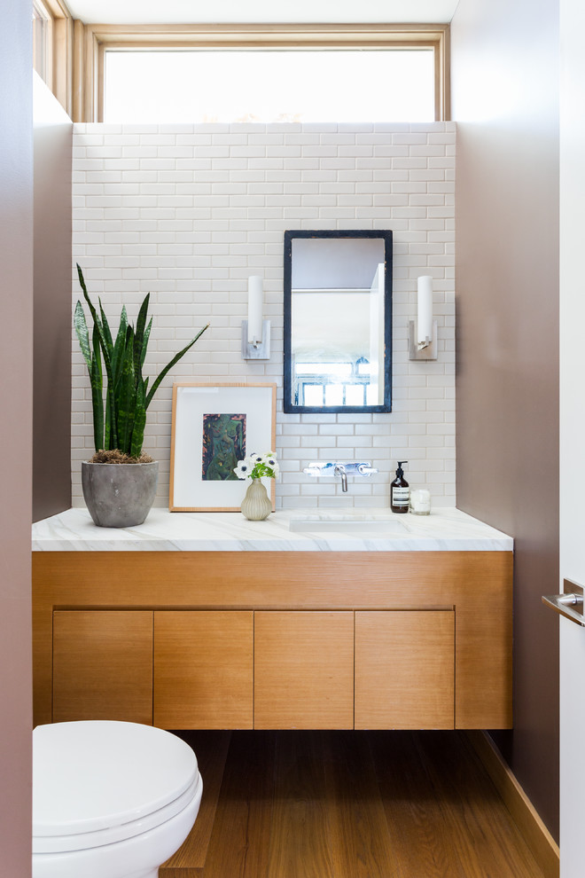 Inspiration för ett mellanstort funkis vit vitt toalett, med släta luckor, skåp i mellenmörkt trä, vit kakel, tunnelbanekakel, bruna väggar, mellanmörkt trägolv, ett undermonterad handfat, marmorbänkskiva och brunt golv