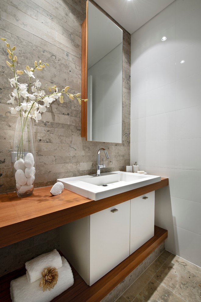 Foto på ett litet funkis brun toalett, med ett nedsänkt handfat, släta luckor, vita skåp, träbänkskiva, vita väggar, kalkstensgolv och kakelplattor