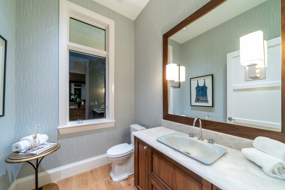 Inspiration for a mid-sized transitional medium tone wood floor and beige floor powder room remodel in Miami with recessed-panel cabinets, medium tone wood cabinets, a two-piece toilet, gray walls, a drop-in sink and gray countertops