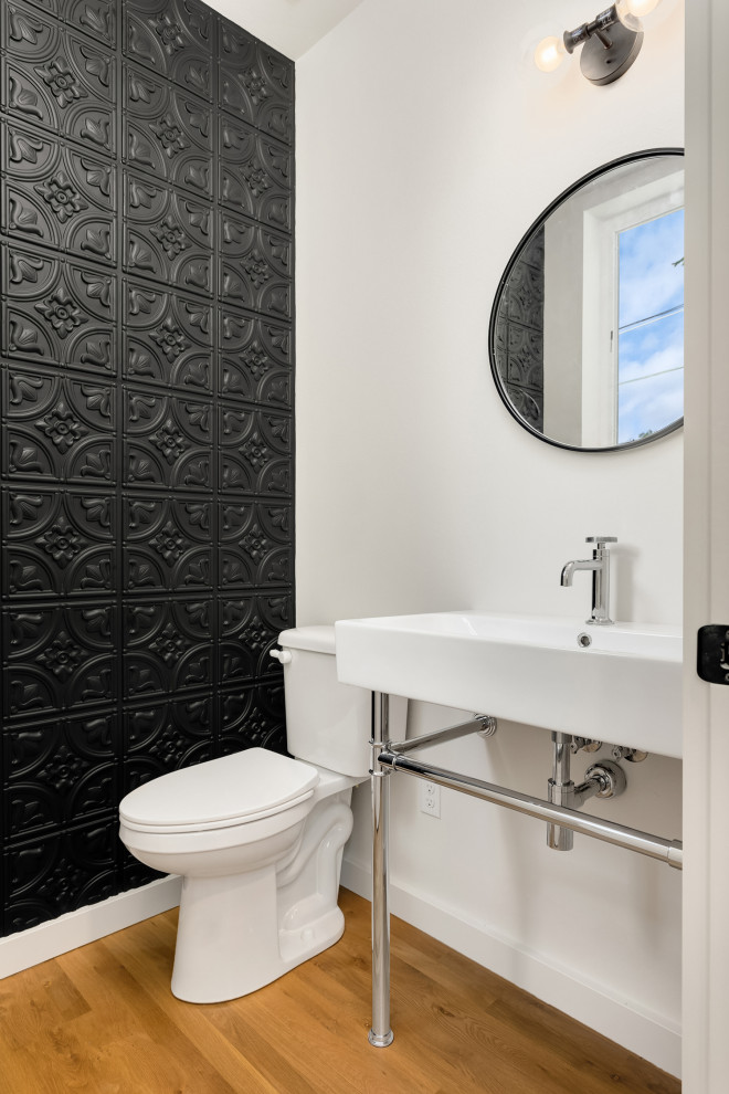 Bild på ett industriellt toalett, med en toalettstol med separat cisternkåpa, svart kakel, vita väggar, mellanmörkt trägolv, ett konsol handfat och brunt golv