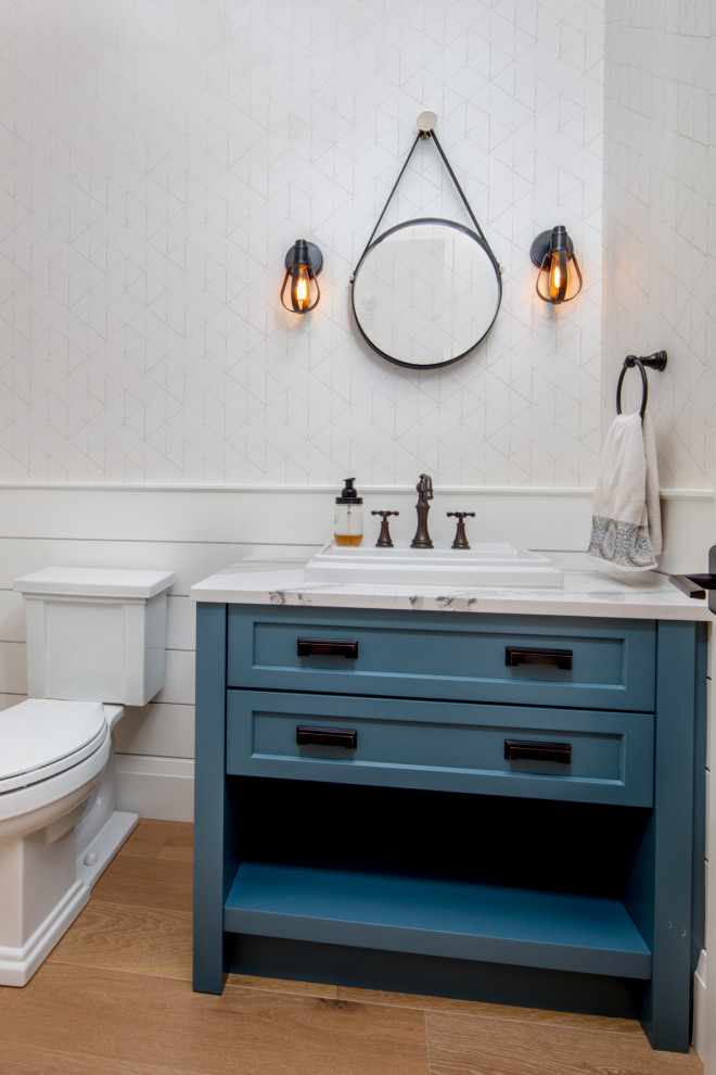 Example of a mid-sized transitional light wood floor, shiplap wall, wainscoting and wallpaper powder room design in Toronto with shaker cabinets, blue cabinets, a two-piece toilet, white walls, multicolored countertops and a freestanding vanity
