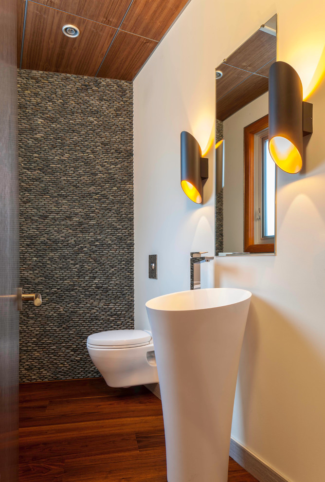 Moderne Gästetoilette mit Sockelwaschbecken, Wandtoilette, grauen Fliesen, Kieselfliesen, weißer Wandfarbe und dunklem Holzboden in Sonstige