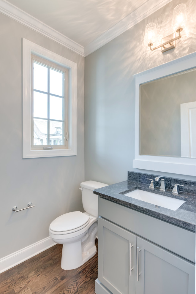 Inredning av ett amerikanskt grå grått toalett, med luckor med infälld panel, grå skåp, en toalettstol med separat cisternkåpa, grå väggar, mörkt trägolv, ett nedsänkt handfat, granitbänkskiva och brunt golv