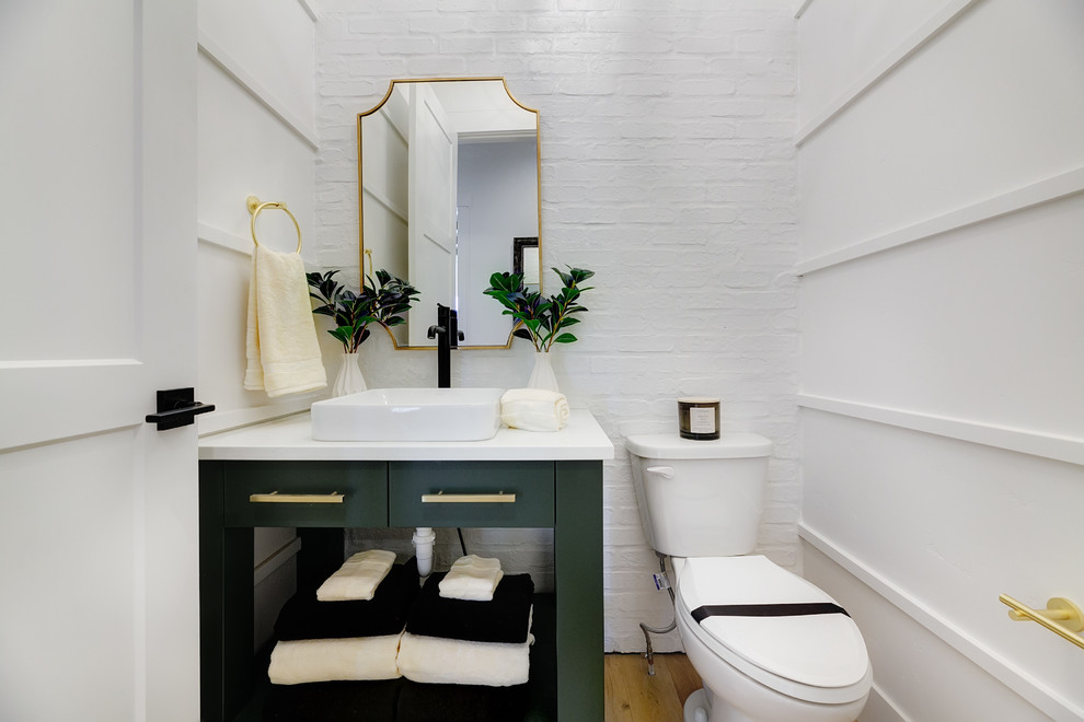 Inredning av ett lantligt mellanstort vit vitt toalett, med möbel-liknande, gröna skåp, en toalettstol med separat cisternkåpa, vita väggar, ljust trägolv, ett fristående handfat, bänkskiva i akrylsten och brunt golv