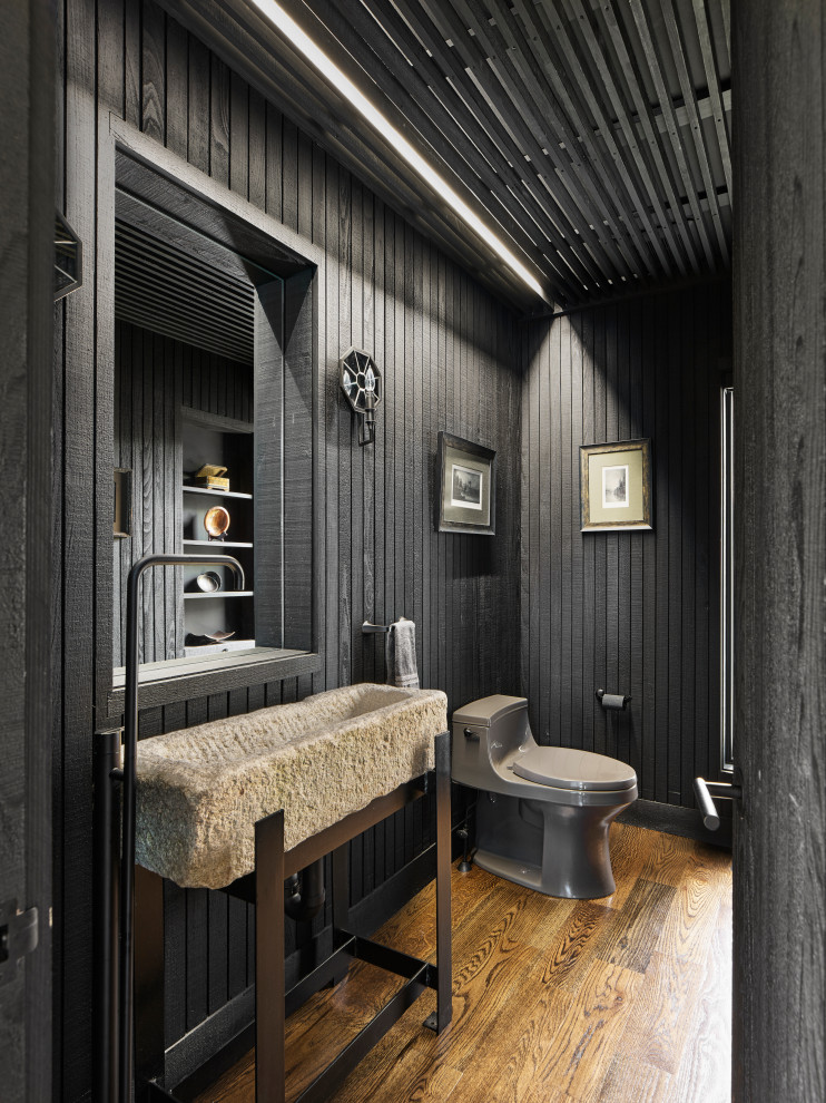 Example of a cottage medium tone wood floor, brown floor and wall paneling powder room design in Philadelphia with gray cabinets, a one-piece toilet, limestone countertops, a freestanding vanity, black walls and a console sink