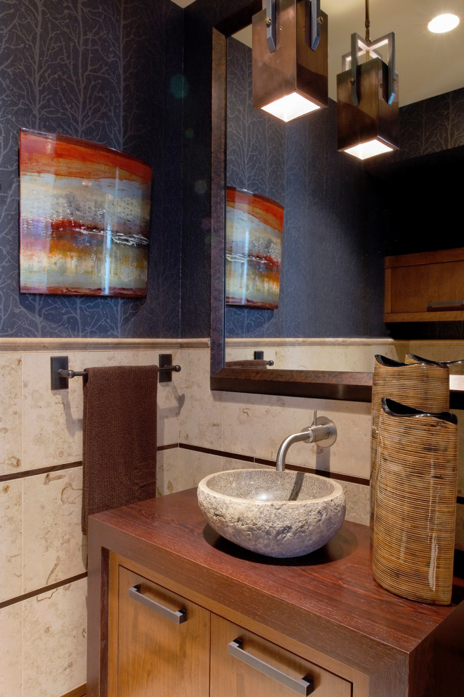 Cette image montre un WC et toilettes design en bois brun de taille moyenne avec une vasque, un placard à porte plane, un plan de toilette en bois, un carrelage beige, un carrelage de pierre et un mur bleu.