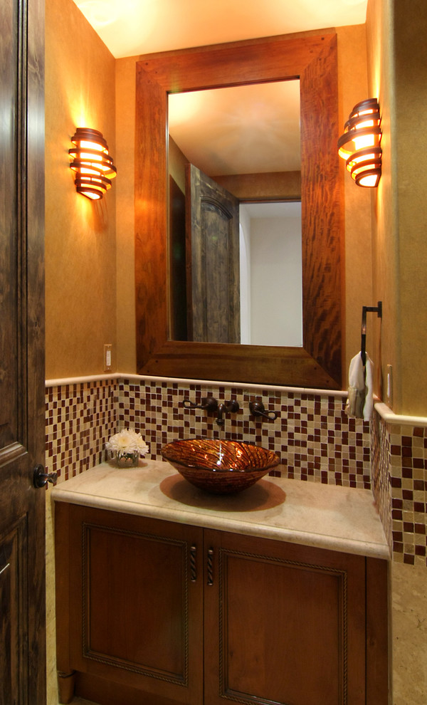 Powder room - mid-sized mediterranean multicolored tile and mosaic tile powder room idea in Los Angeles with recessed-panel cabinets, medium tone wood cabinets, a one-piece toilet, beige walls, a vessel sink and limestone countertops