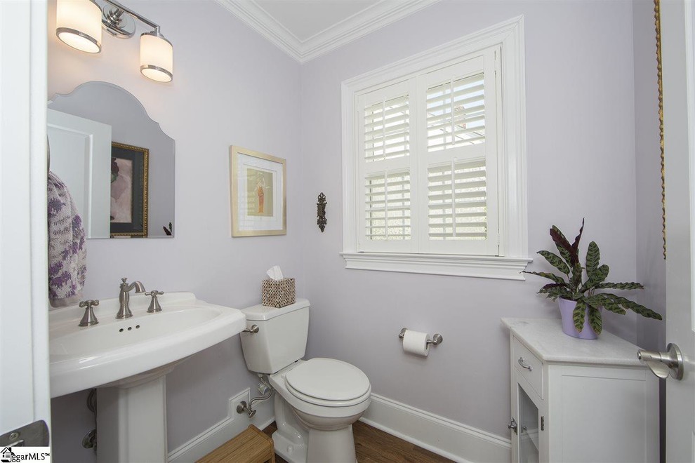Bild på ett litet vintage toalett, med en toalettstol med hel cisternkåpa, lila väggar, mörkt trägolv, ett piedestal handfat och brunt golv