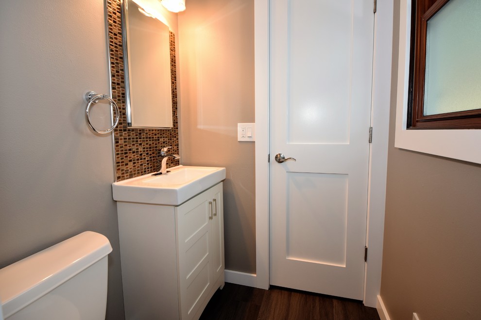 Foto på ett litet amerikanskt toalett, med skåp i shakerstil, vita skåp, en toalettstol med separat cisternkåpa, mosaik, grå väggar, mörkt trägolv, ett integrerad handfat och brunt golv