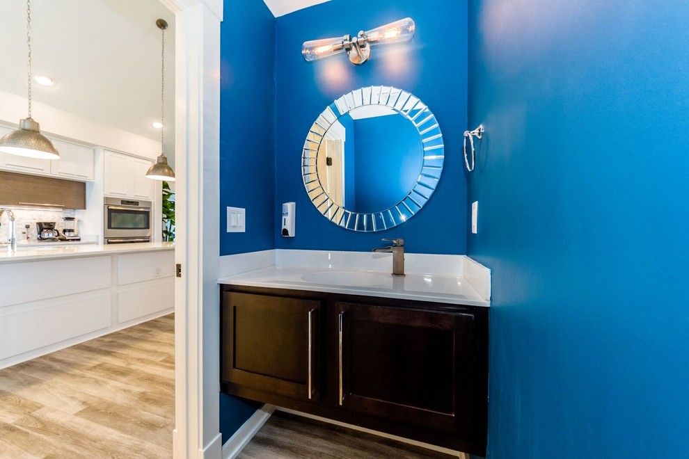 Ejemplo de aseo costero pequeño con armarios estilo shaker, puertas de armario de madera en tonos medios, paredes azules, suelo de madera clara, lavabo integrado, encimera de acrílico y suelo marrón