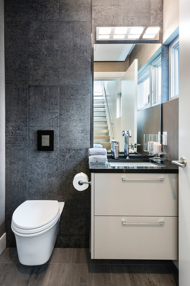 Small modern cloakroom in Vancouver with flat-panel cabinets, white cabinets, a wall mounted toilet, grey walls, porcelain flooring, a submerged sink, brown floors and black worktops.