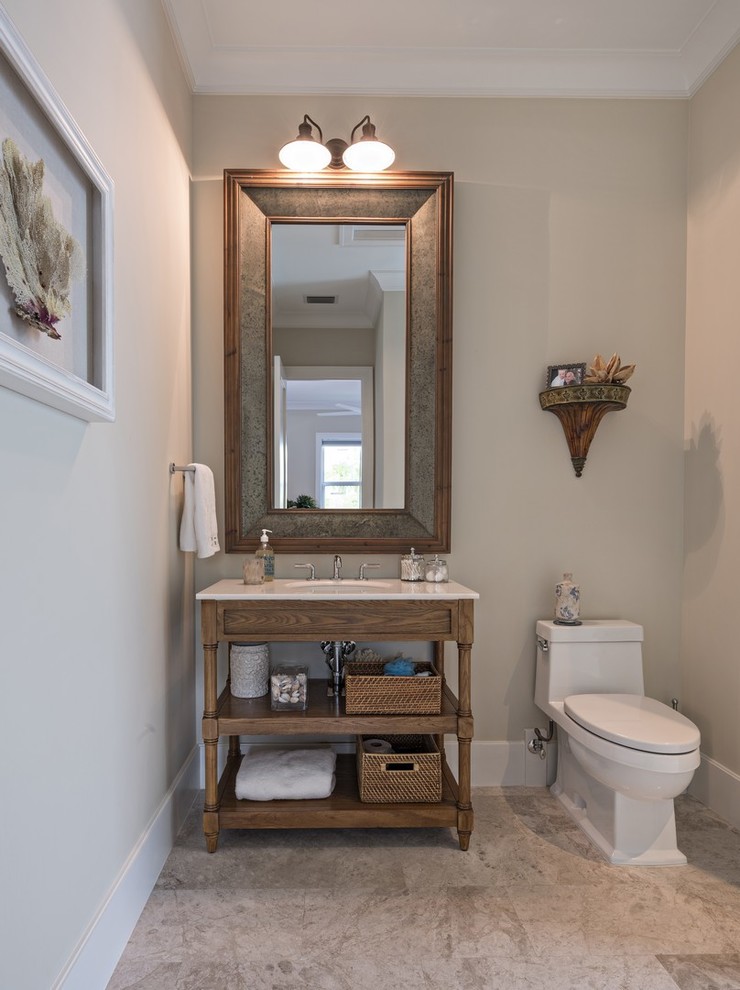 This is an example of a traditional cloakroom in Miami with freestanding cabinets, medium wood cabinets, a one-piece toilet, beige walls, marble flooring, a submerged sink, marble worktops, beige floors, grey tiles and white worktops.