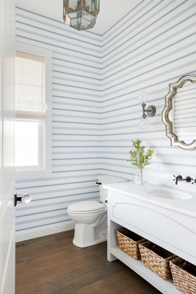 Cette image montre un grand WC et toilettes marin avec un placard en trompe-l'oeil, des portes de placard blanches, WC à poser, un mur bleu, un lavabo encastré, un plan de toilette en marbre, un sol marron, un sol en bois brun et un plan de toilette blanc.