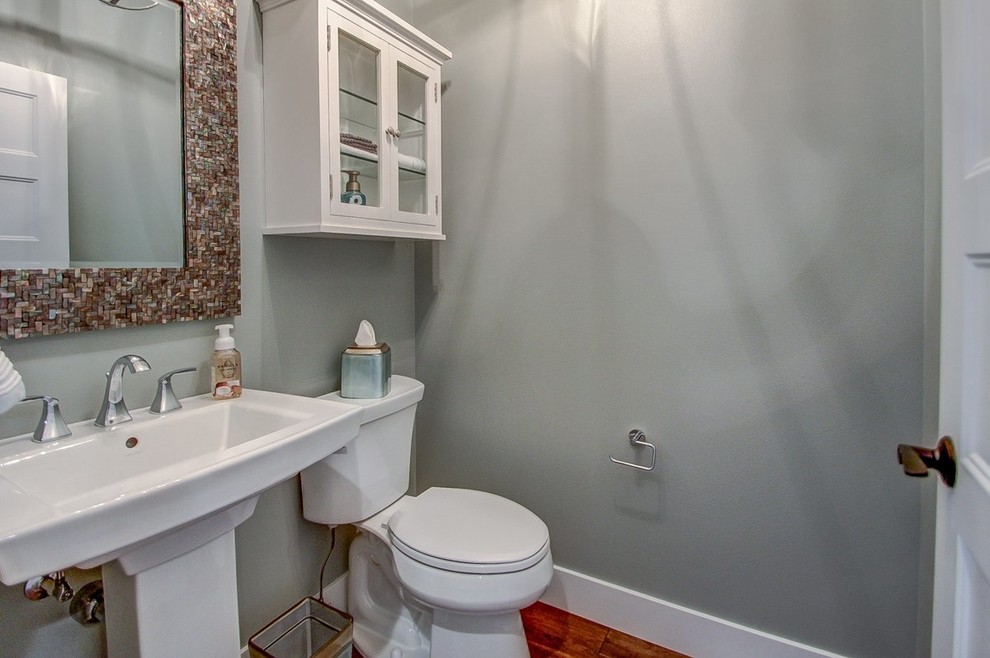 Klassisk inredning av ett litet toalett, med luckor med glaspanel, vita skåp, en toalettstol med separat cisternkåpa, grå väggar, mellanmörkt trägolv och ett piedestal handfat