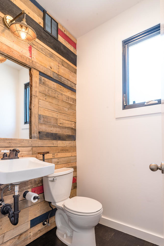 Industriell inredning av ett toalett, med mörkt trägolv, ett väggmonterat handfat, en toalettstol med separat cisternkåpa, flerfärgade väggar och brunt golv