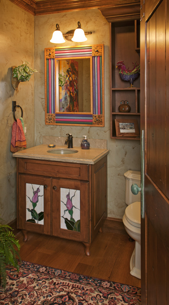 Bild på ett litet rustikt toalett, med luckor med glaspanel, en toalettstol med separat cisternkåpa, beige väggar, mörkt trägolv, ett undermonterad handfat, bänkskiva i travertin och brunt golv