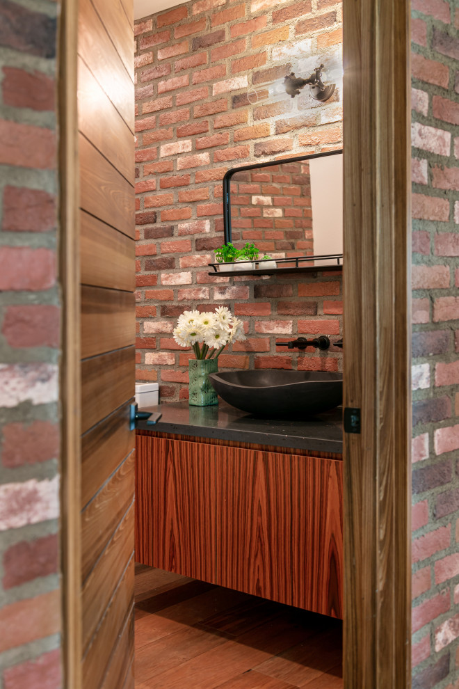 Modelo de aseo flotante actual con armarios con paneles lisos, puertas de armario de madera oscura, paredes rojas, suelo de madera oscura, lavabo sobreencimera, suelo marrón, encimeras negras y ladrillo