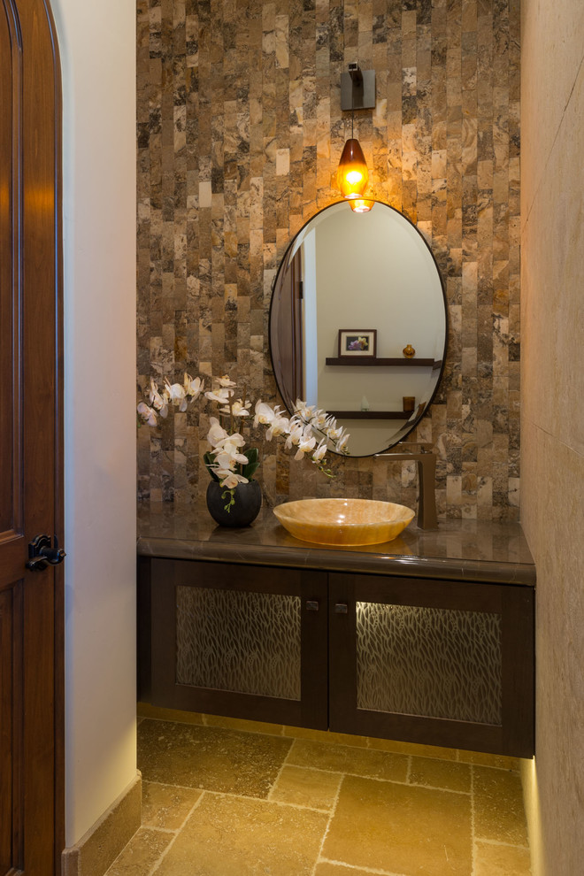 This is an example of a small mediterranean cloakroom in San Diego with glass-front cabinets, dark wood cabinets, beige tiles, stone tiles, multi-coloured walls, limestone flooring, a vessel sink, marble worktops and beige floors.