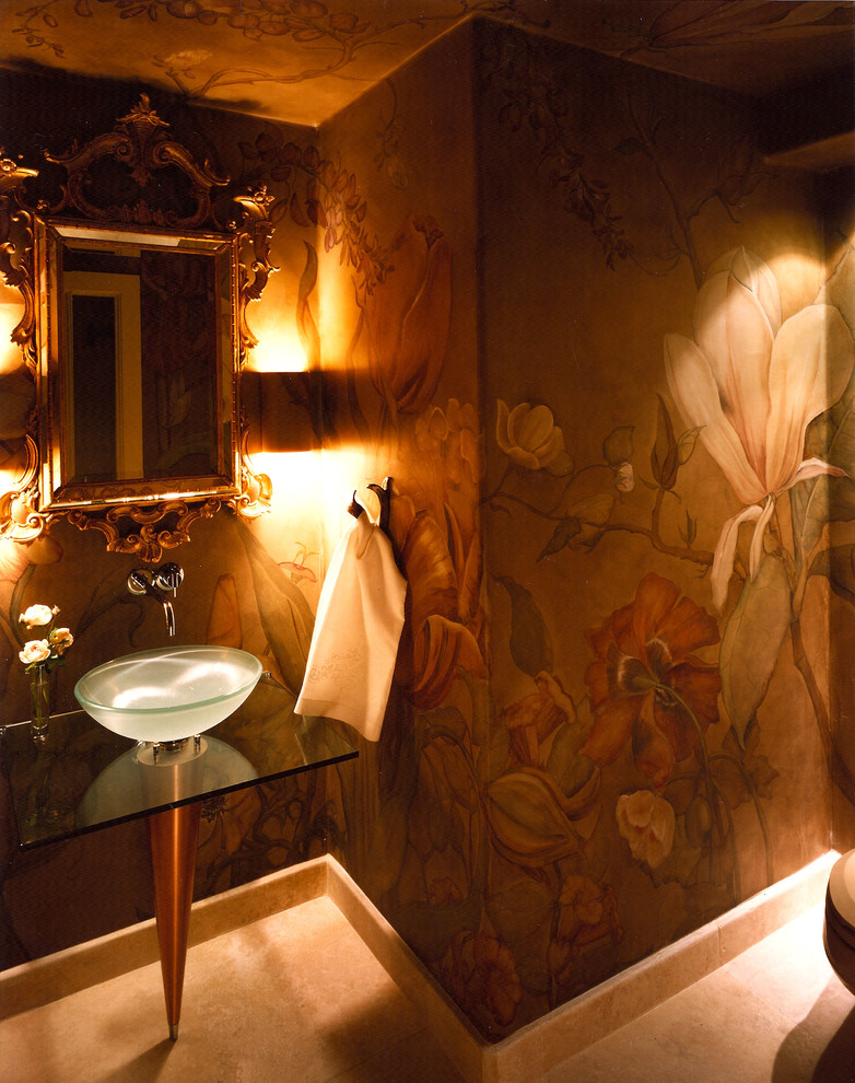 Cette image montre un WC et toilettes design avec une vasque, un plan de toilette en verre, WC à poser, un carrelage beige, un carrelage de pierre, un mur multicolore et un sol en calcaire.