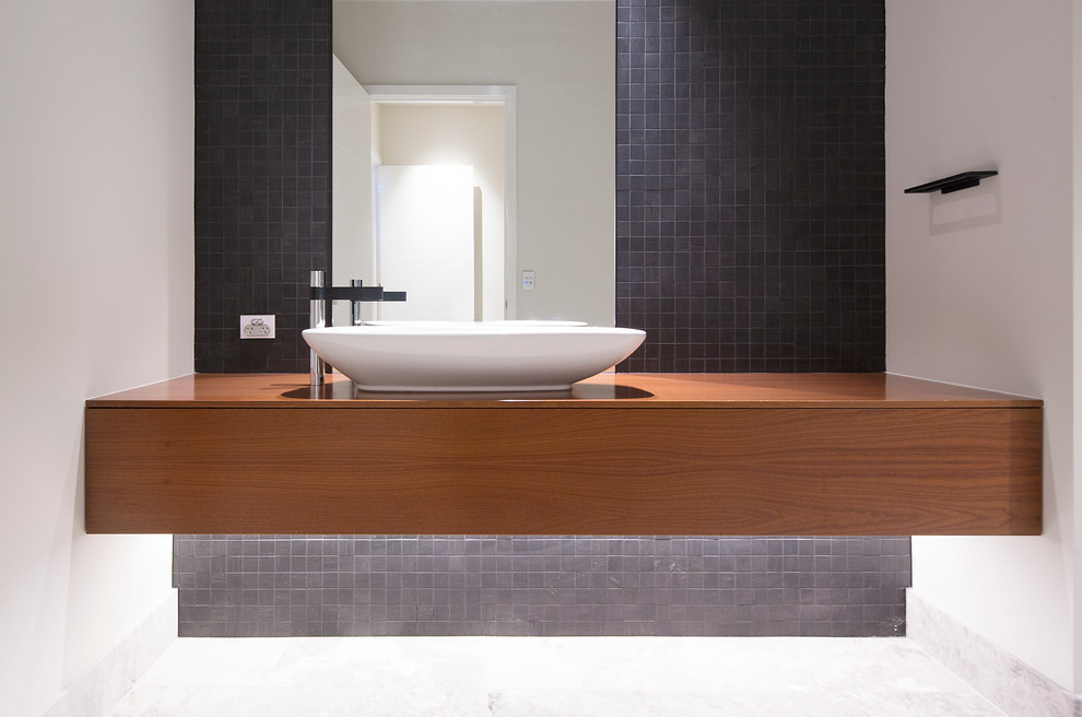 Cette photo montre un petit WC et toilettes moderne en bois brun avec un placard à porte plane, un carrelage noir, des carreaux de céramique, un mur blanc, un sol en marbre, une vasque, un plan de toilette en bois et un plan de toilette marron.
