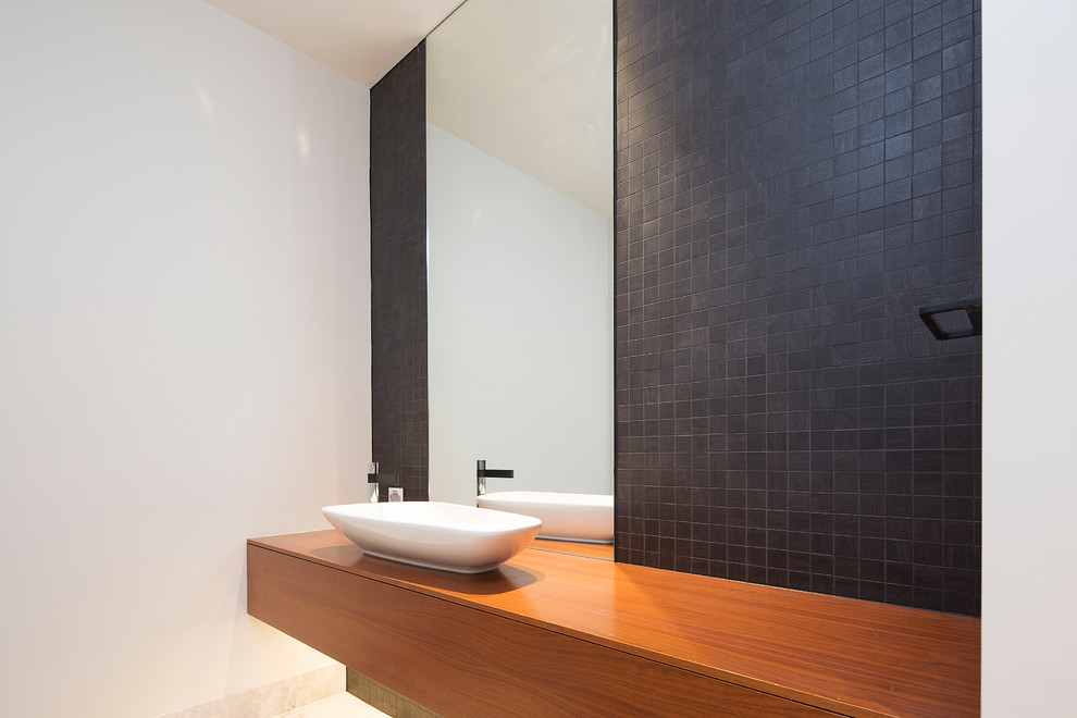 Cette image montre un petit WC et toilettes minimaliste en bois brun avec un placard à porte plane, un carrelage noir, des carreaux de céramique, un mur blanc, un sol en marbre, une vasque, un plan de toilette en bois et un plan de toilette marron.