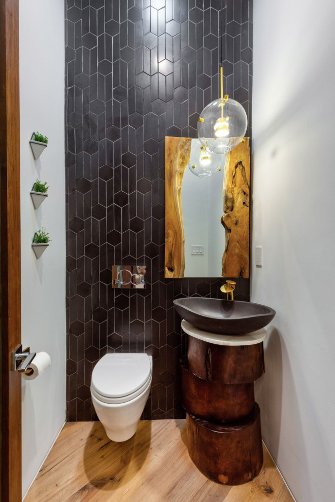 Imagen de aseo actual con sanitario de pared, baldosas y/o azulejos negros, paredes blancas, suelo de madera clara, lavabo sobreencimera y suelo beige