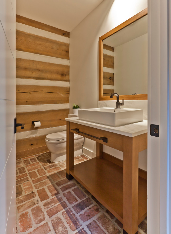 Mid-sized danish brick floor powder room photo in Indianapolis with furniture-like cabinets, medium tone wood cabinets, a one-piece toilet, white walls, a vessel sink and marble countertops
