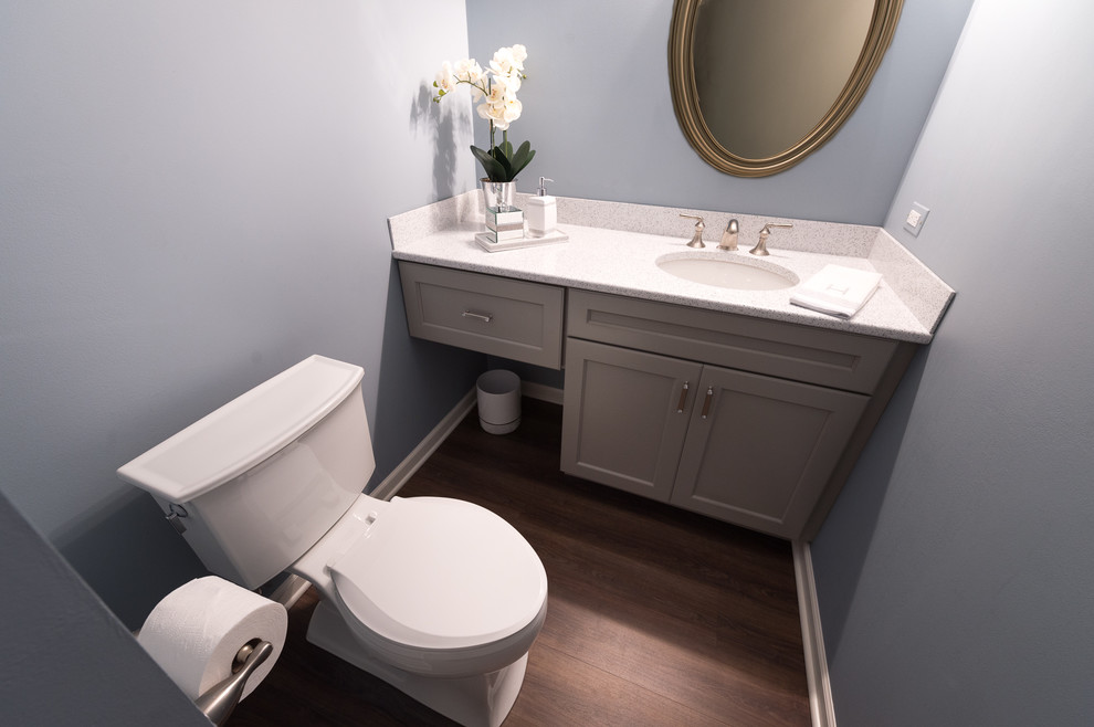 Bild på ett litet vintage toalett, med luckor med infälld panel, grå skåp, en toalettstol med separat cisternkåpa, blå väggar, vinylgolv, ett undermonterad handfat, bänkskiva i kvarts, brunt golv och stenhäll