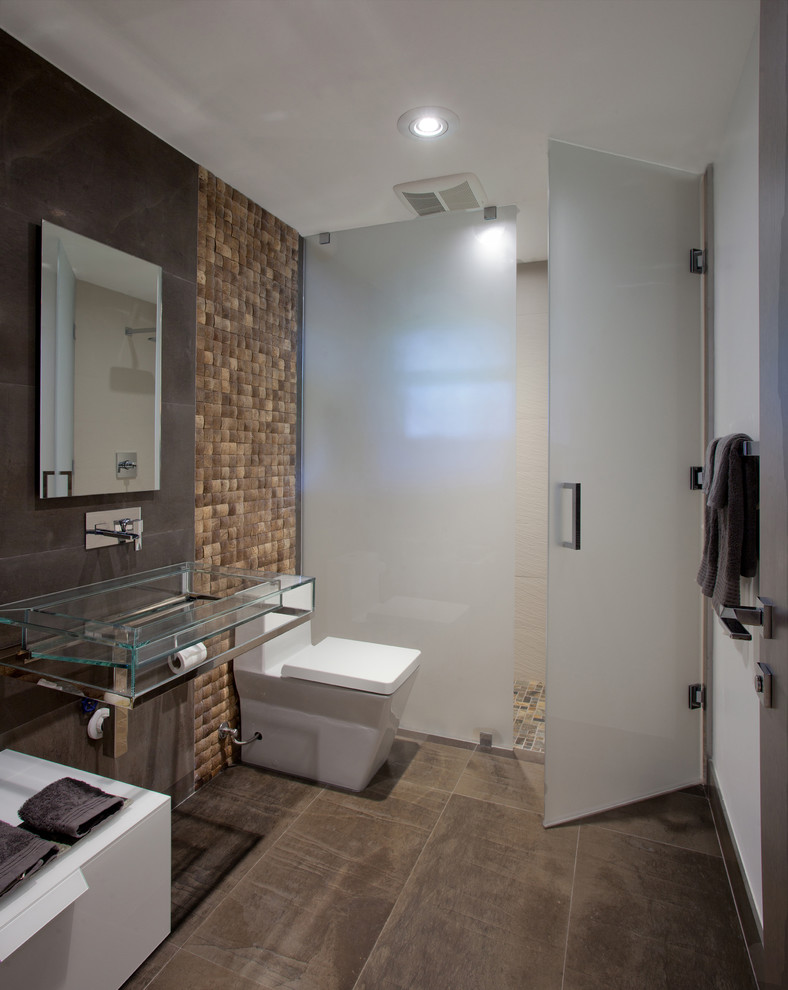 This is an example of a small contemporary cloakroom in Miami with a one-piece toilet, beige tiles, brown tiles, stone tiles, brown walls, ceramic flooring, a pedestal sink, glass worktops and beige floors.