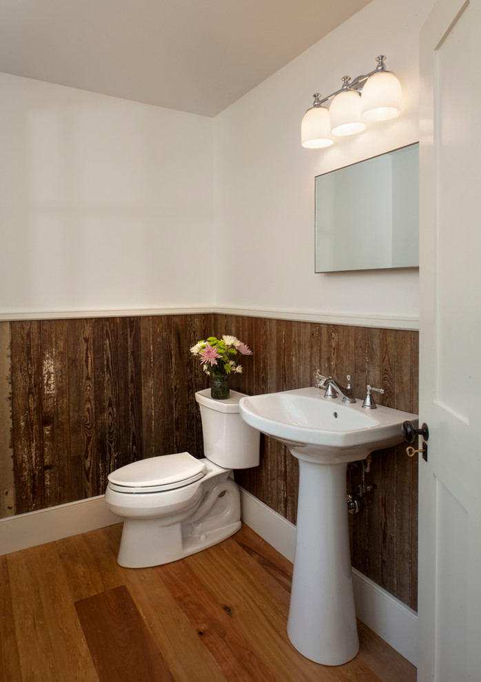 Modelo de aseo de estilo de casa de campo de tamaño medio con lavabo con pedestal, sanitario de dos piezas, paredes blancas y suelo de madera en tonos medios