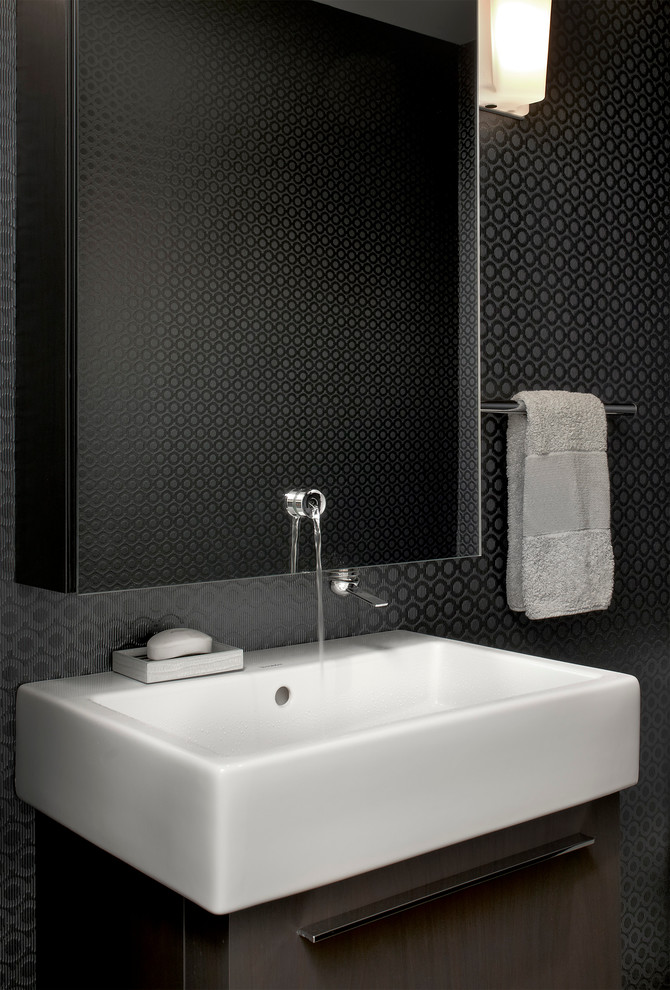 Bild på ett litet funkis toalett, med ett väggmonterat handfat, släta luckor, skåp i mörkt trä och svarta väggar