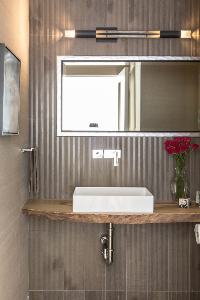 Bild på ett litet funkis brun brunt toalett, med en vägghängd toalettstol, grå kakel, stenkakel, grå väggar, ljust trägolv, ett fristående handfat, träbänkskiva och grått golv