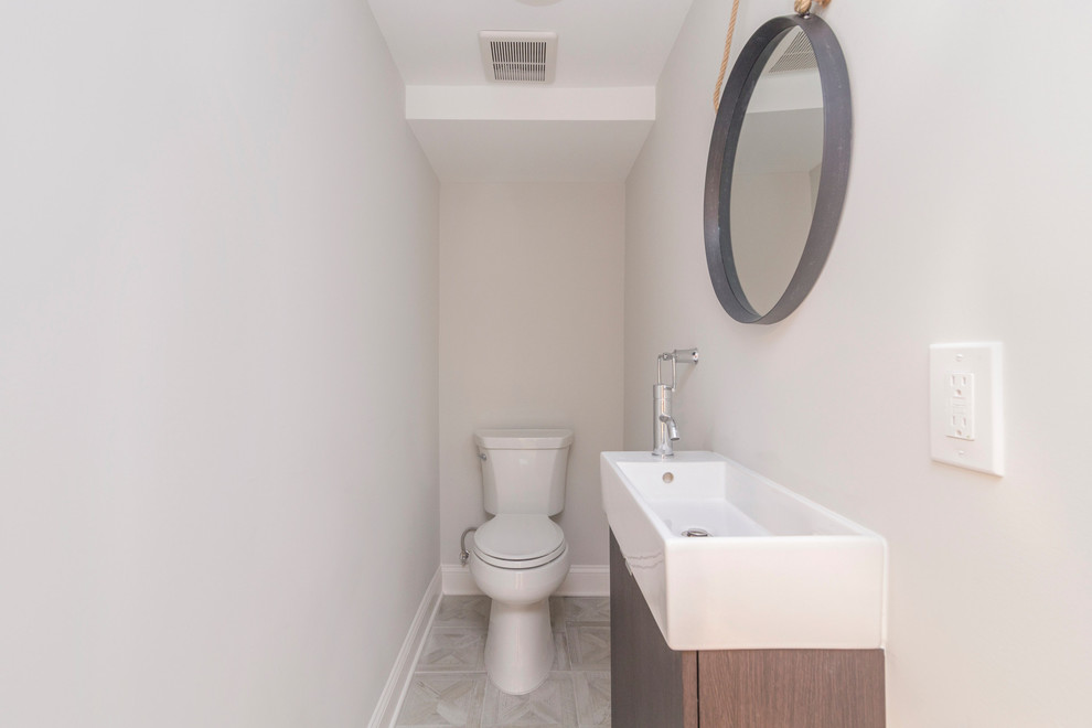 This is an example of a small modern cloakroom in New York with flat-panel cabinets, medium wood cabinets, a one-piece toilet, grey walls, a trough sink and beige floors.