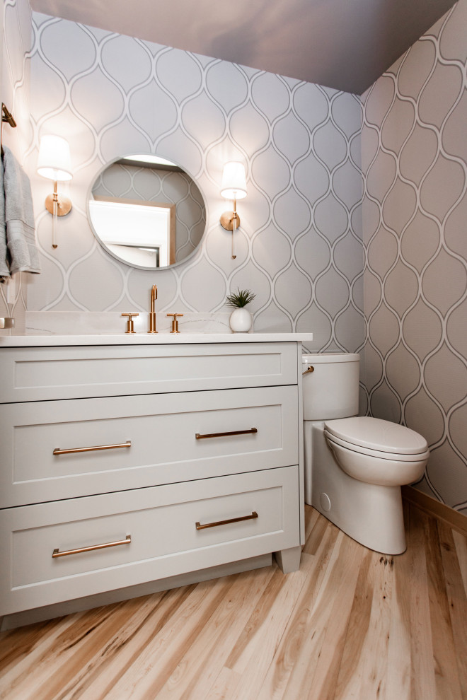 Example of a small medium tone wood floor, brown floor and wallpaper powder room design in Minneapolis with shaker cabinets, gray cabinets, a one-piece toilet, gray walls, an integrated sink, quartz countertops, white countertops and a freestanding vanity