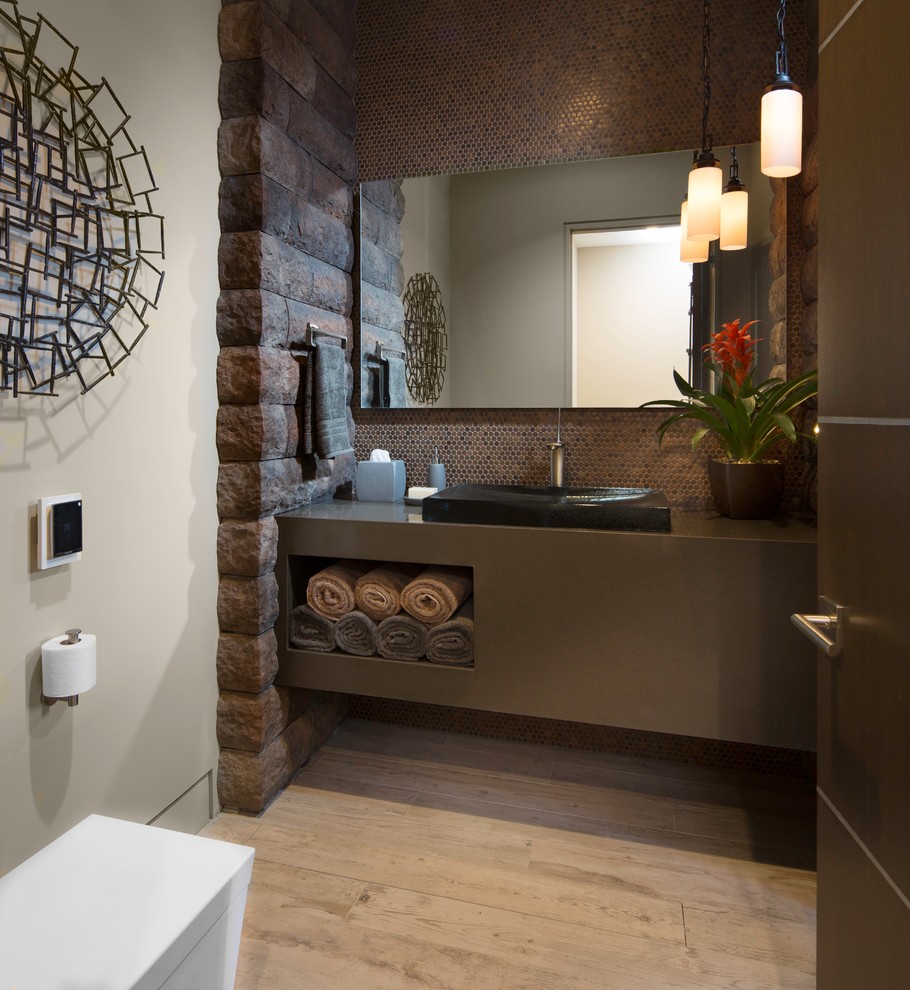 Cette image montre un WC et toilettes design de taille moyenne avec des portes de placard grises, un carrelage marron, mosaïque, un mur gris, parquet clair, une vasque, un plan de toilette en stratifié, un placard sans porte et un sol beige.