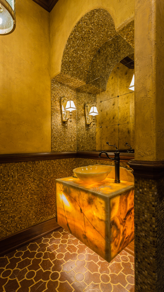 На фото: туалет в средиземноморском стиле с разноцветной плиткой, керамической плиткой, бежевыми стенами, полом из терракотовой плитки, настольной раковиной, столешницей из оникса и коричневым полом с