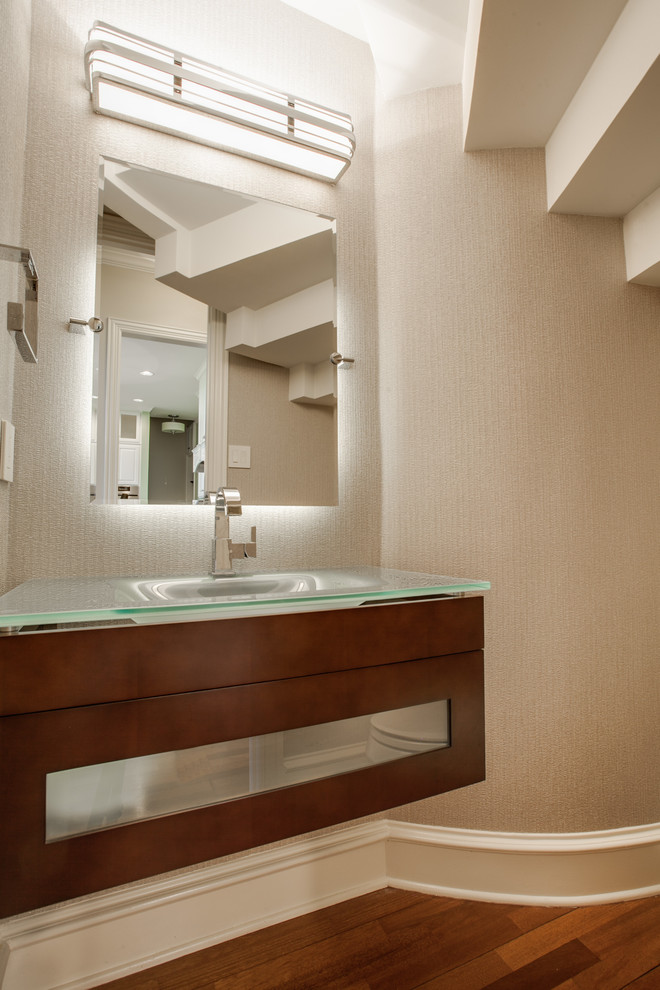 Cette photo montre un petit WC suspendu tendance en bois foncé avec un lavabo intégré, un placard à porte vitrée, un plan de toilette en verre, un mur beige et un sol en bois brun.